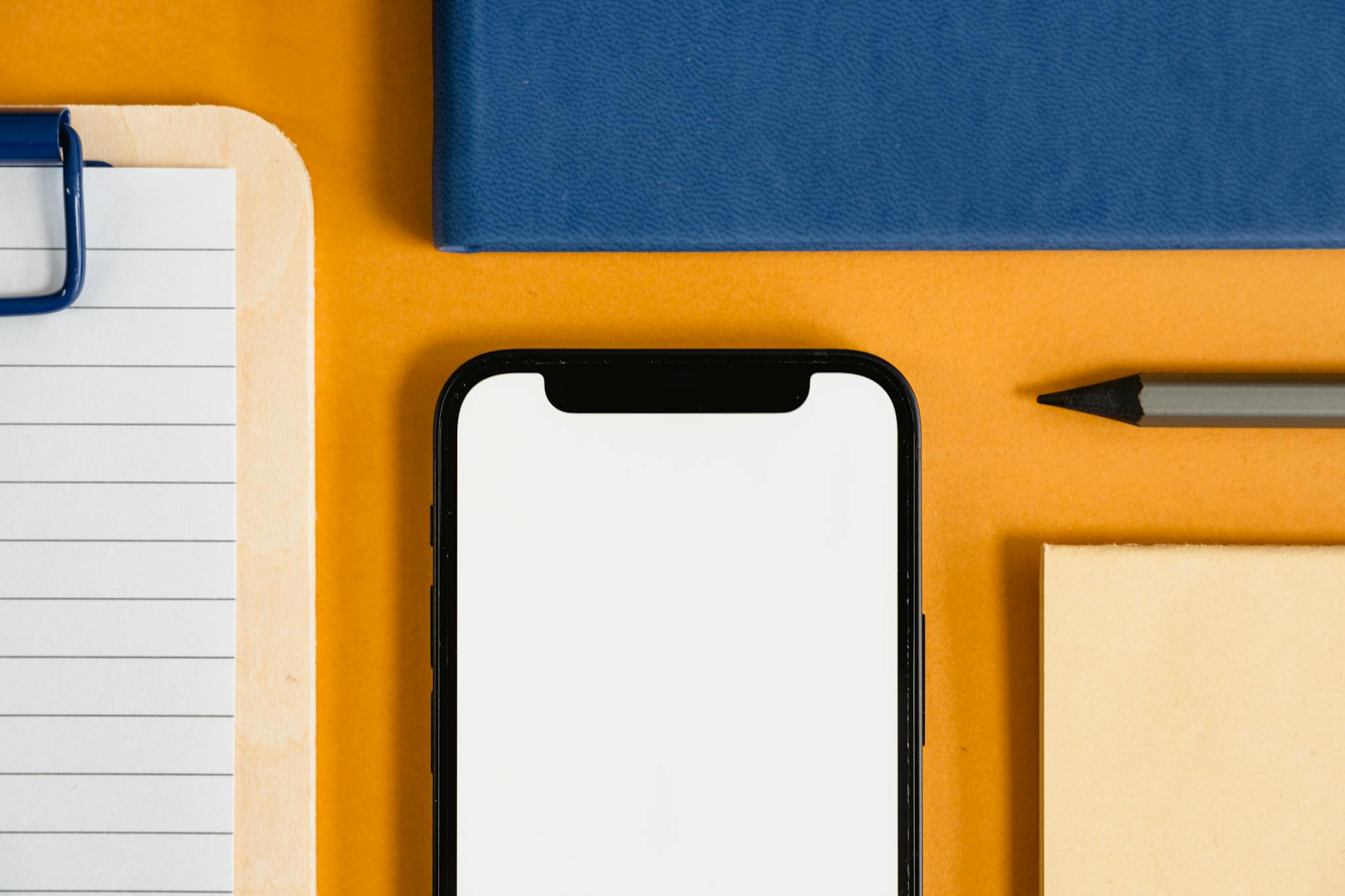 A flat lay of office supplies, including a smartphone, clipboard, pencil, on a vibrant orange desk.